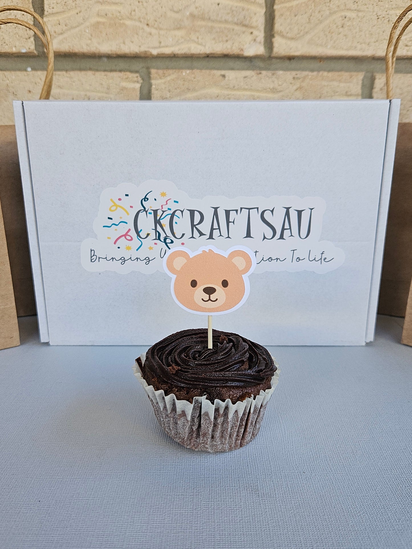 Cupcake with a bear topper next to a branded box on a textured surface.
