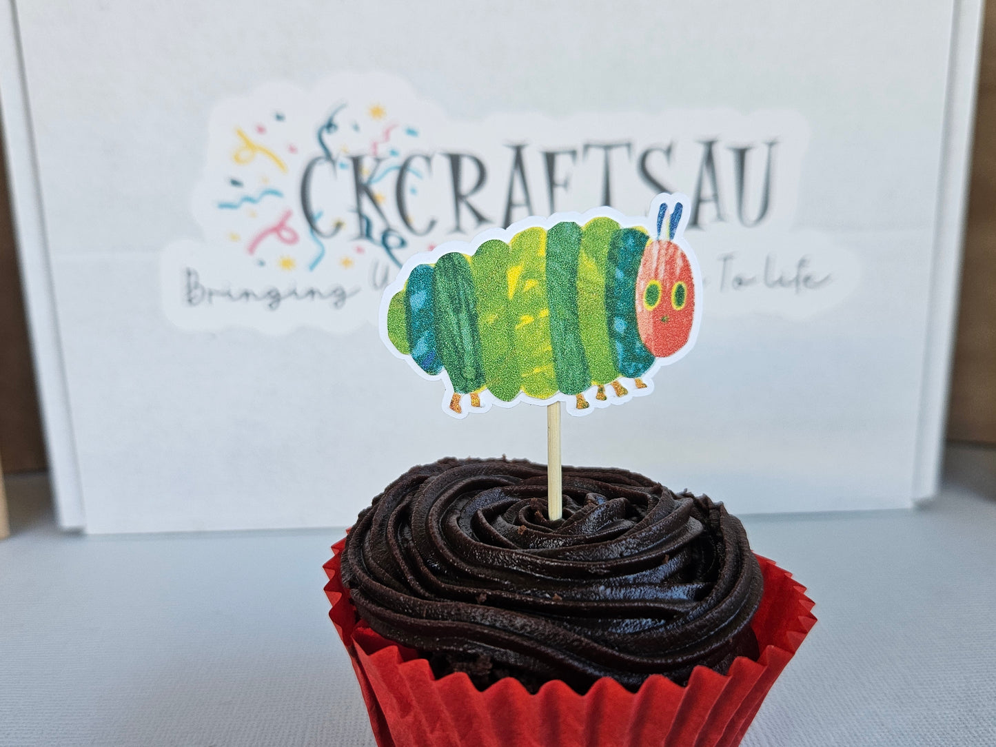 Cupcake with a caterpillar topper in front of a sign.