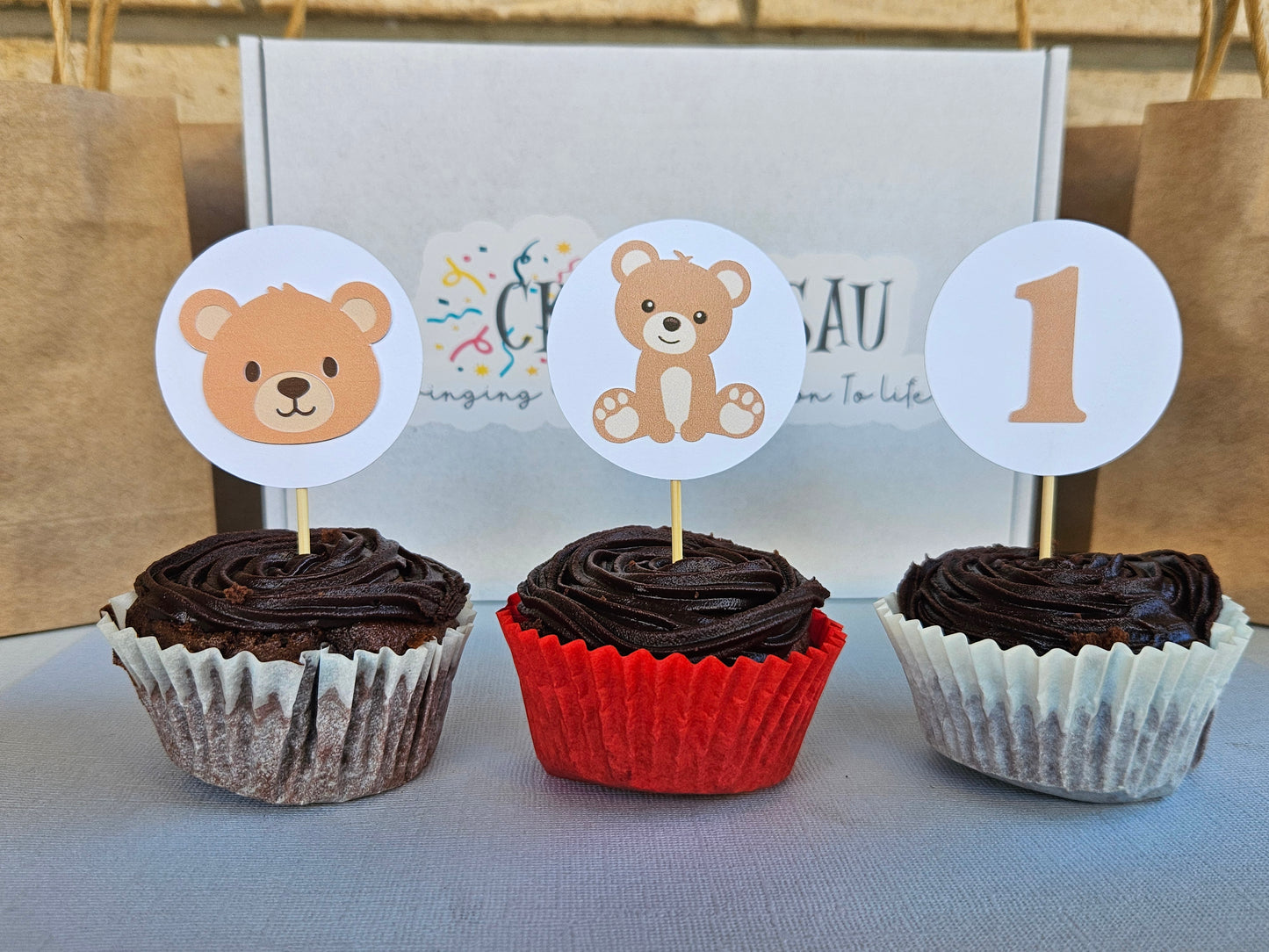 Three cupcakes with chocolate frosting and bear-themed toppers on a light background.
