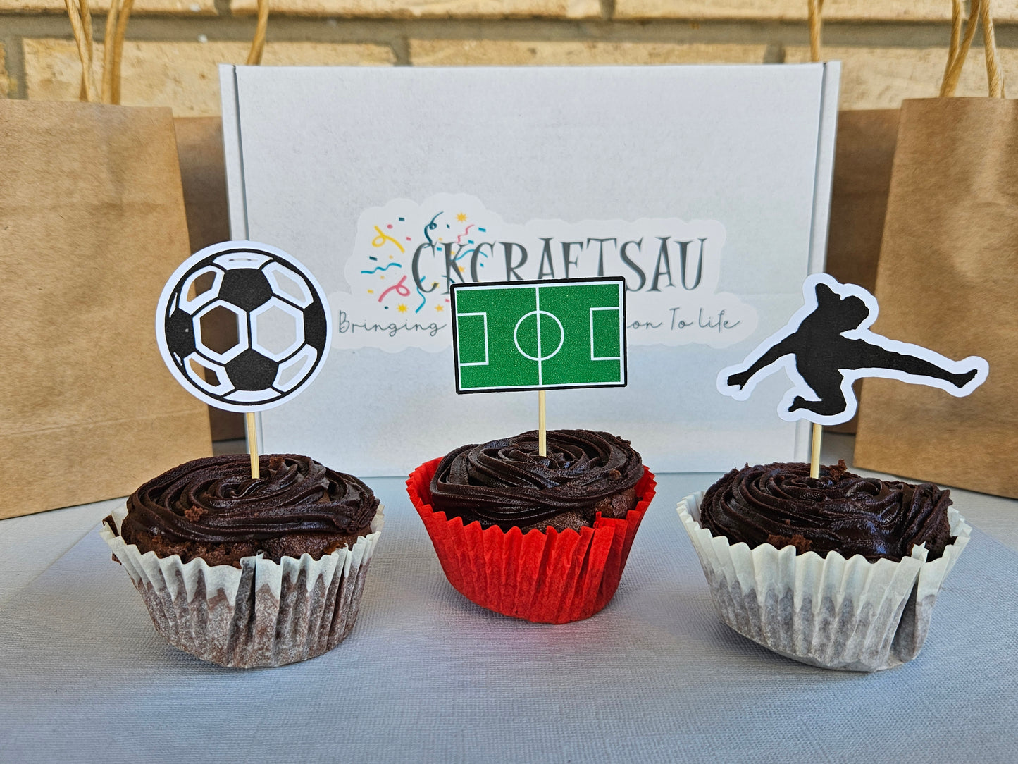 Three chocolate cupcakes with sports-themed toppers on a table with a branded box in the background.