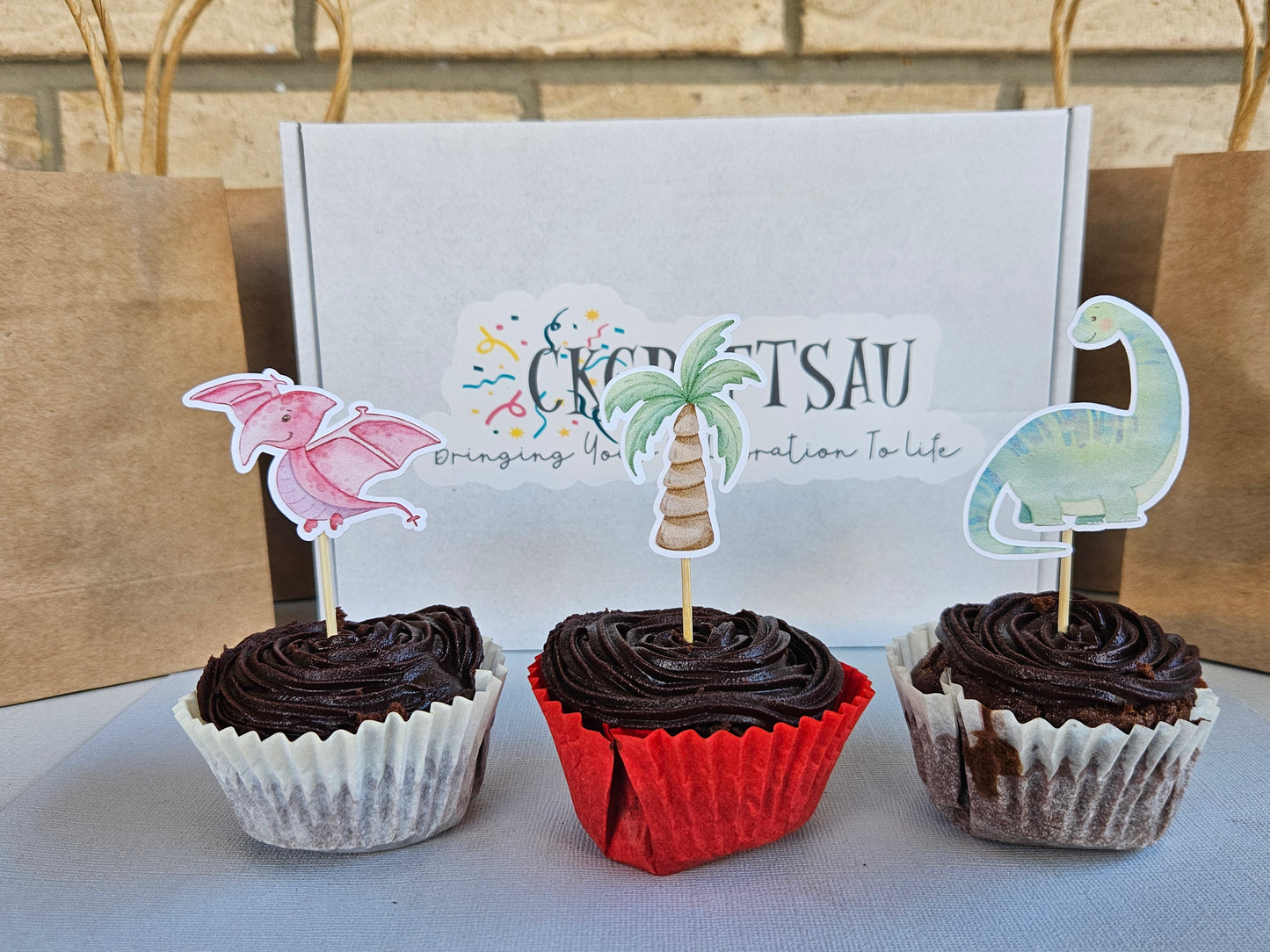 Three chocolate cupcakes with dino toppers in front of a branded box.