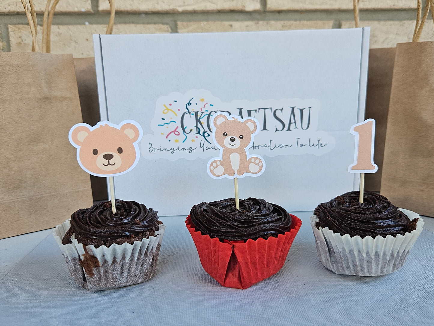 Three cupcakes with chocolate frosting and bear-themed toppers in front of a branded box.