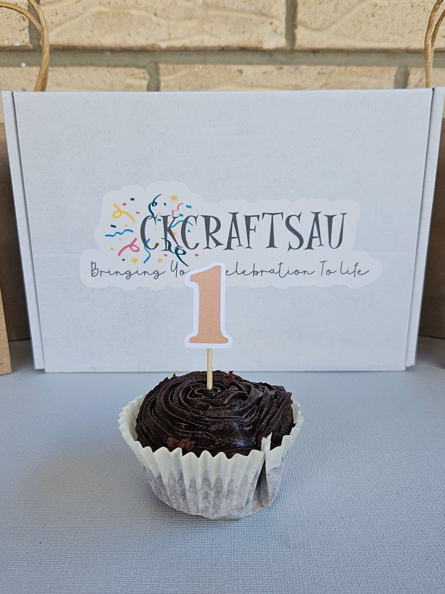 Chocolate cupcake with a number 1 topper next to a white box with branding.