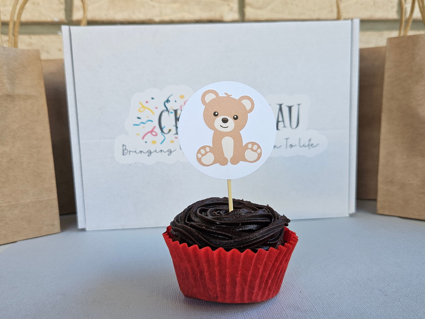 Chocolate cupcake with a teddy bear topper in front of a white box with text and logos.