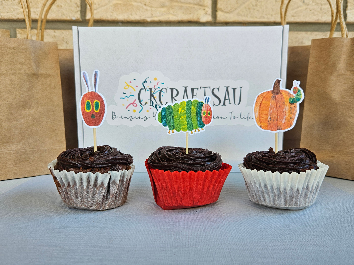 Three cupcakes with chocolate frosting on a gray surface, with a branded box in the background.