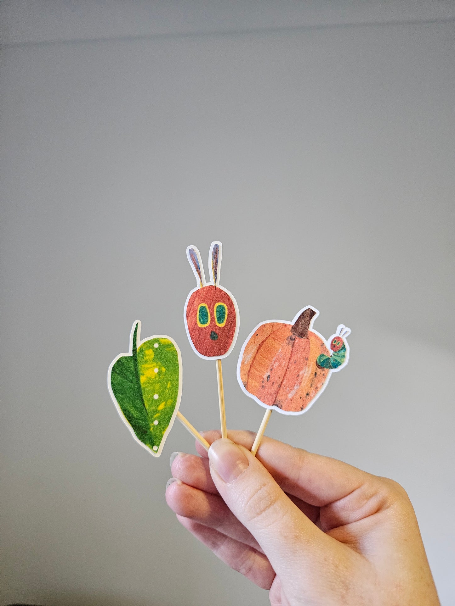 Hand holding a stick with autumn-themed cutouts of a pumpkin, leaf, and caterpillar on a gray background.