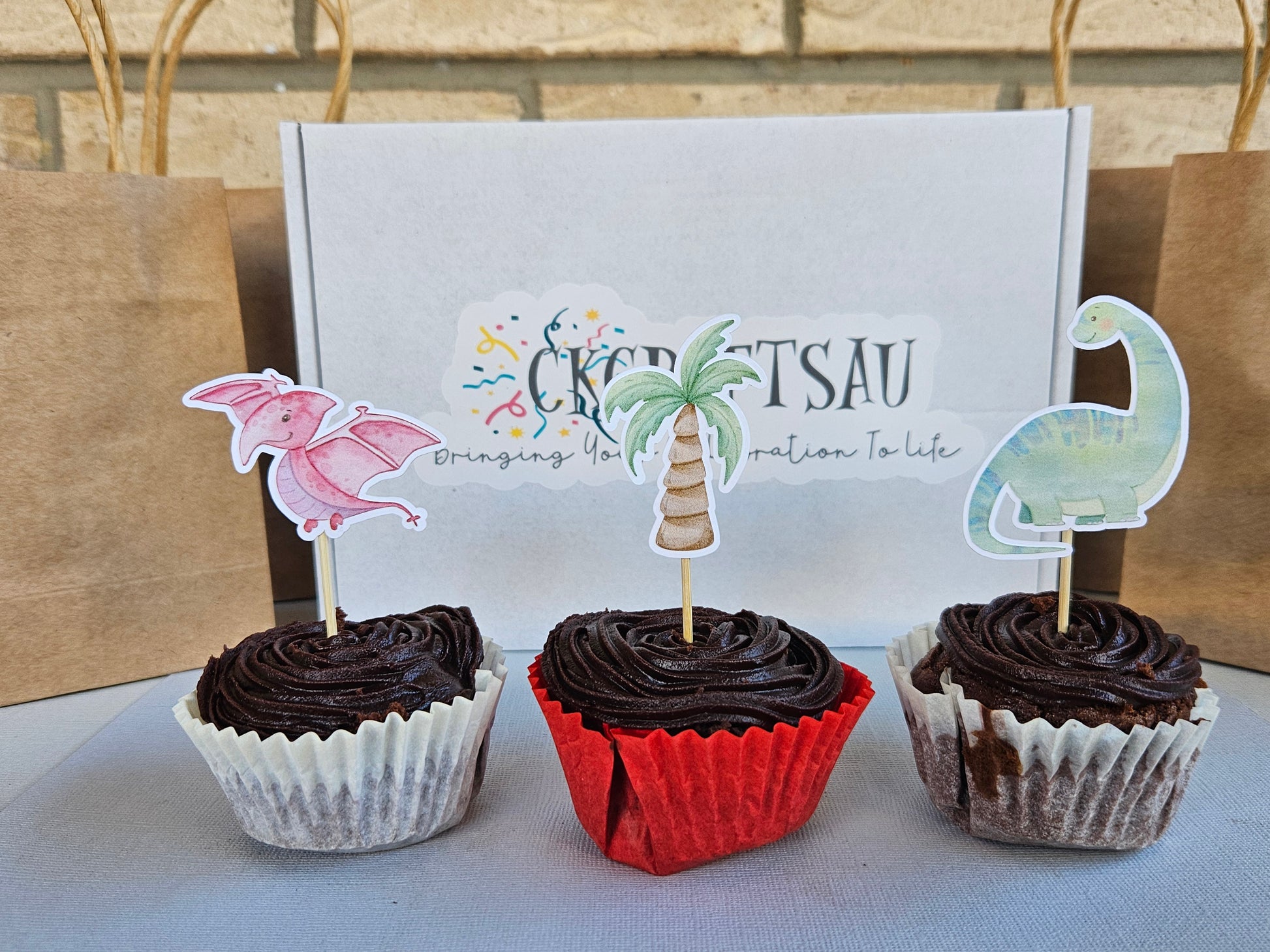 Three chocolate cupcakes with dino toppers in front of a branded box.