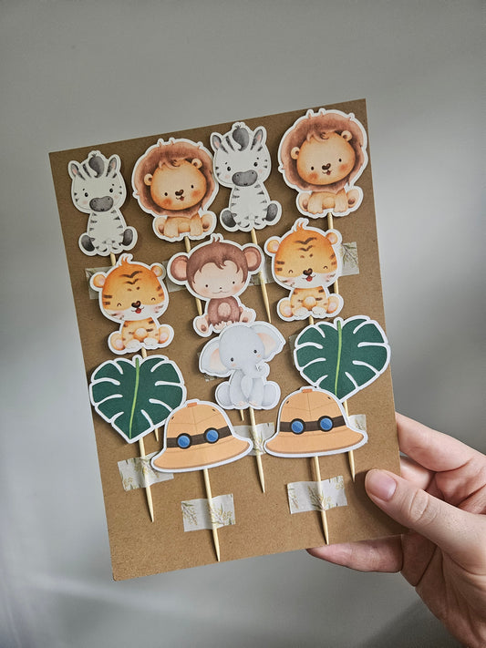 A variety of handcrafted cake toppers including animal figures such as a lion, zebra, elephant, monkey, safari hat, leaf, and tiger, attached to sticks and made from cardstock.