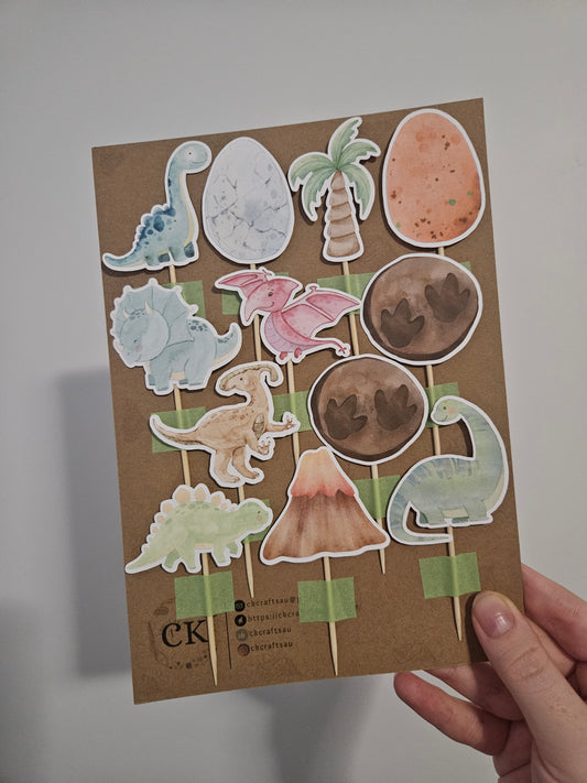 A variety of handcrafted dino cupcake toppers on a bamboo toothpick, in different colors, displayed on a cardstock.