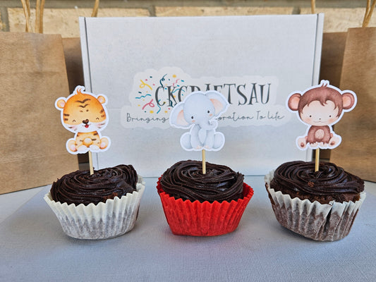 Three cupcakes with animal-themed toppers on a light surface, with a box in the background.