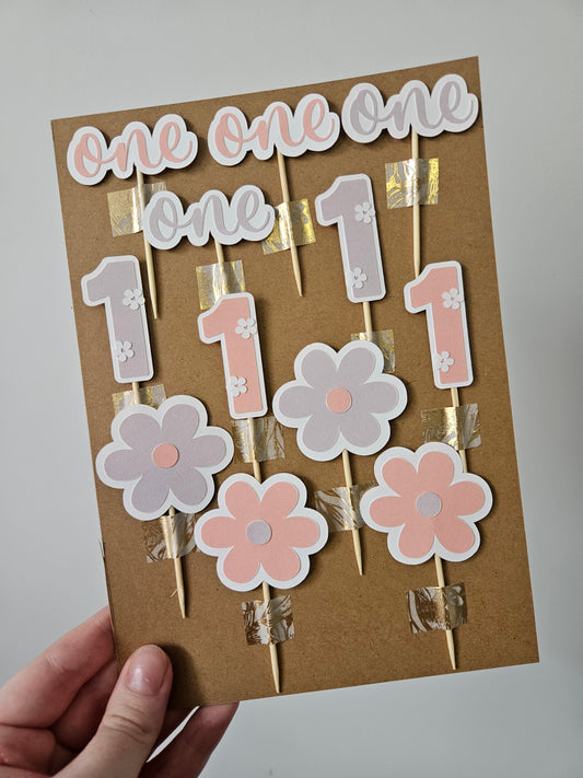 A set of handcrafted floral cake toppers made from cardstock and bamboo toothpicks, featuring numbers and floral designs in pink and purple colors.