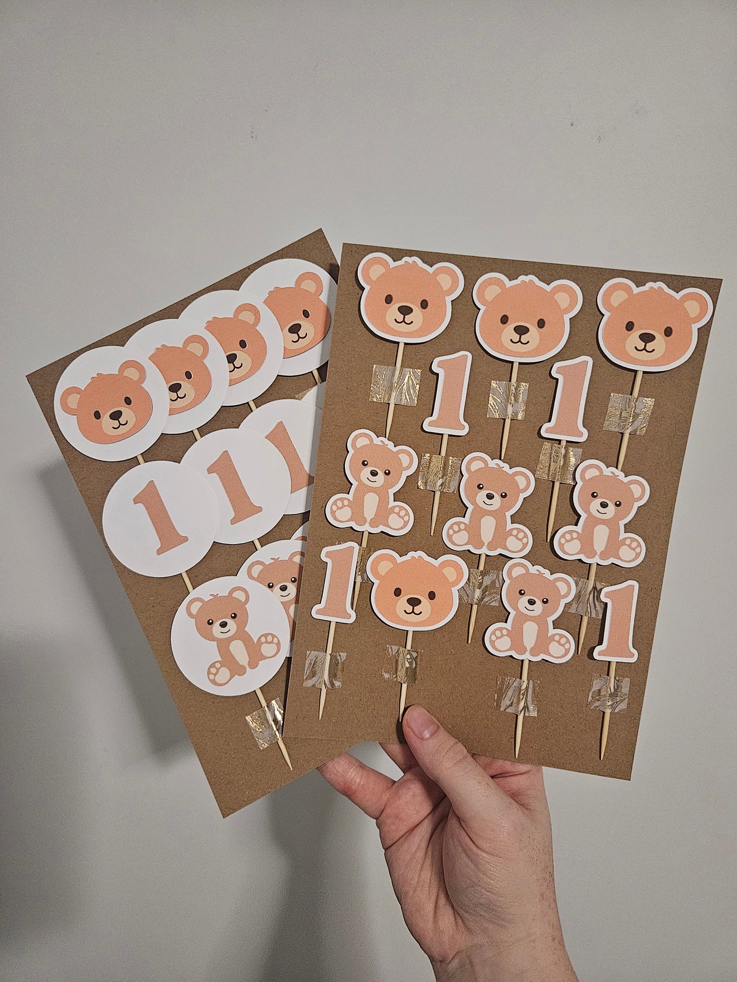Handmade bear cupcake toppers held in a person's hand, with variations including bear heads, numbers, and complete bear designs, predominantly in brown tones.