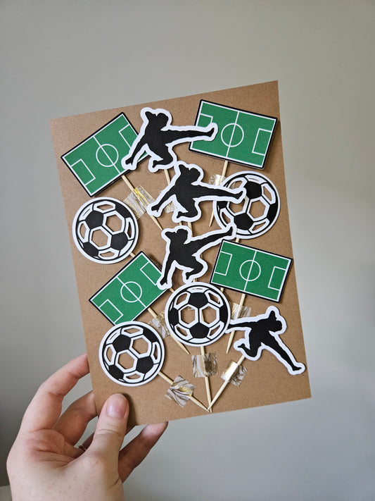 A variety of handmade soccer-themed cake toppers, including silhouettes of soccer players, balls, and fields, held in a person's hand.