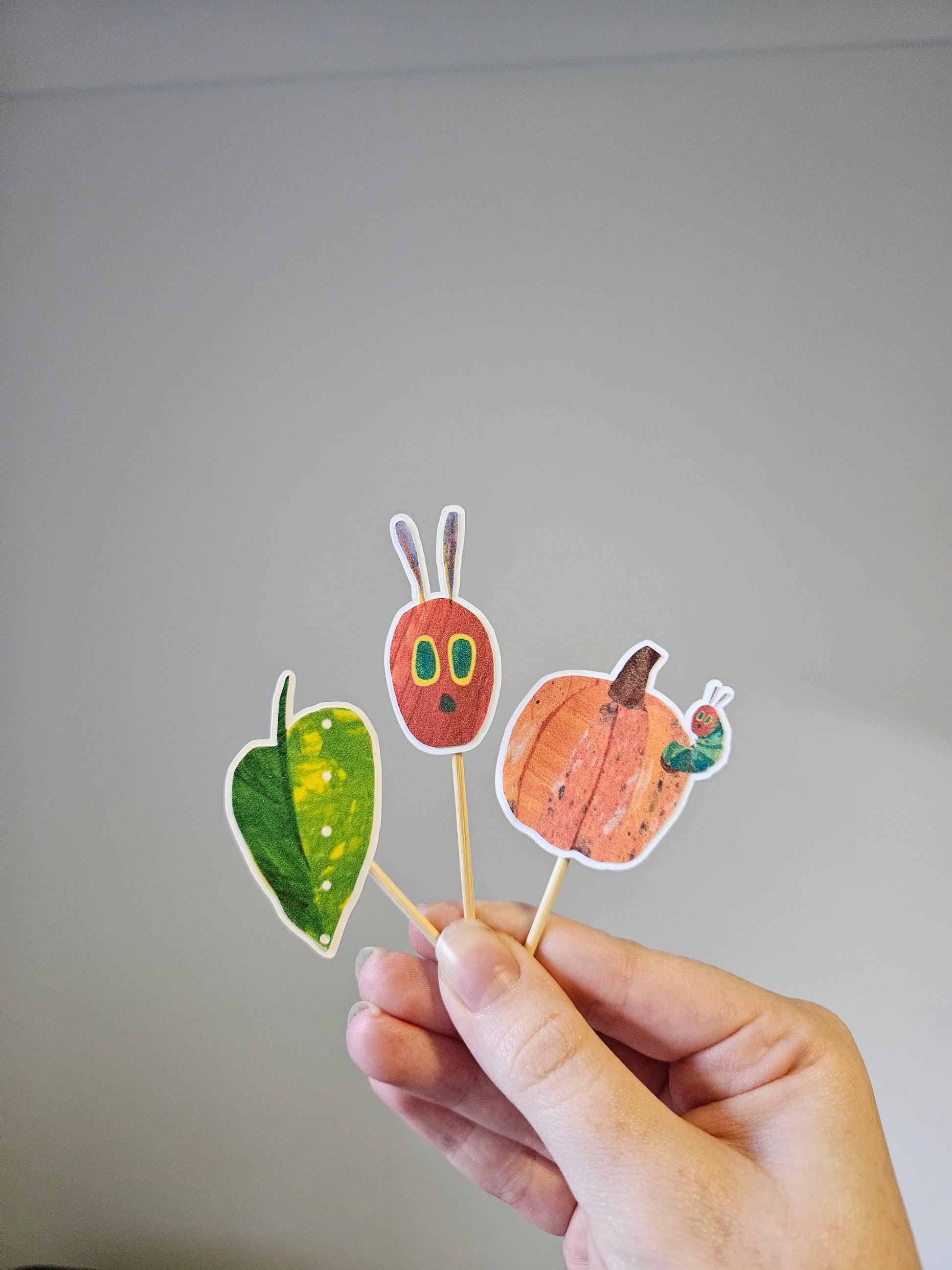 Hand holding a stick with autumn-themed cutouts of a pumpkin, leaf, and caterpillar on a gray background.